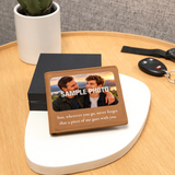 A personalized leather bifold wallet with a printed photo of two men and a sentimental quote, displayed on a white oval stand on a wooden table next to keys and a potted plant.