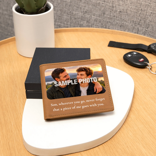 A personalized leather bifold wallet with a printed photo of two men and a sentimental quote, displayed on a white oval stand on a wooden table next to keys and a potted plant.