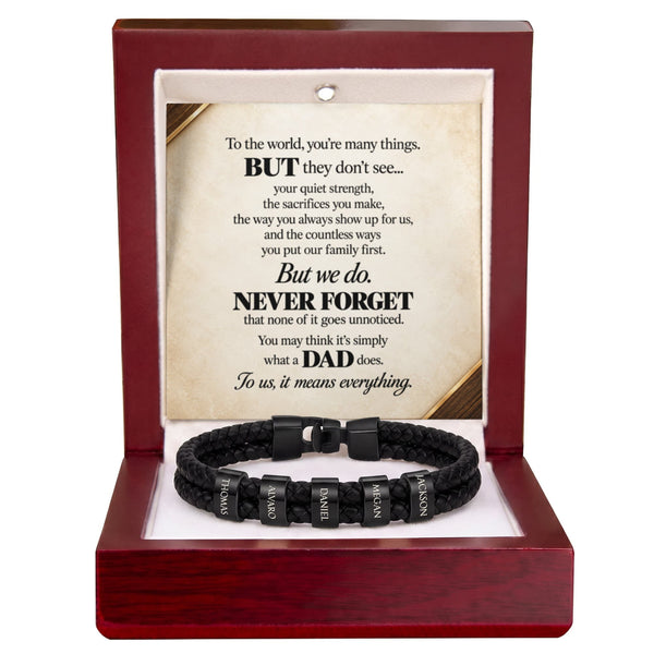 A black braided cord bracelet with silver-toned metal links engraved with names, presented inside an open reddish-brown wooden gift box. The box contains a printed memorial message card addressed to a father.
