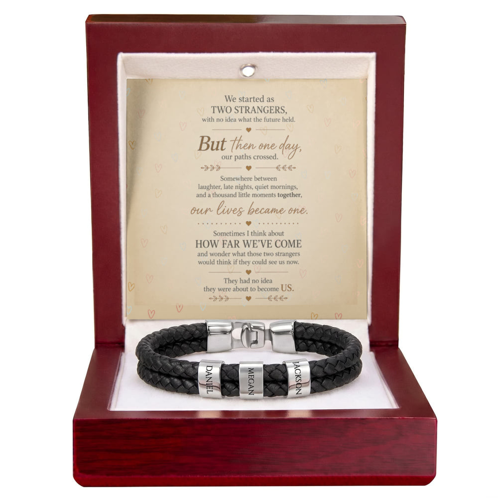 A black braided leather bracelet with silver engraved name plates, presented inside a dark red wooden gift box with a printed romantic message card behind it.