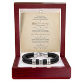 A black braided leather bracelet with silver engraved name plates, presented inside a dark red wooden gift box with a printed romantic message card behind it.