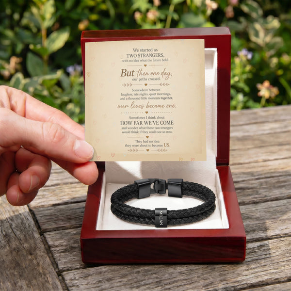A hand holds an open red gift box over a wooden table outdoors, showing a black braided cord bracelet with a black clasp and a printed message card inside the box.