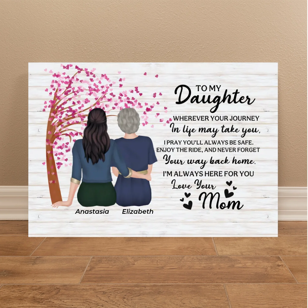 A personalized canvas wall art print leaning against a beige wall on a wooden floor. The print features a white wood-plank background with a black typewriter-style font poem addressed to a daughter, an illustration of two women sitting together under a pink cherry blossom tree, and the names 'Anastasia' and 'Elizabeth' printed below.