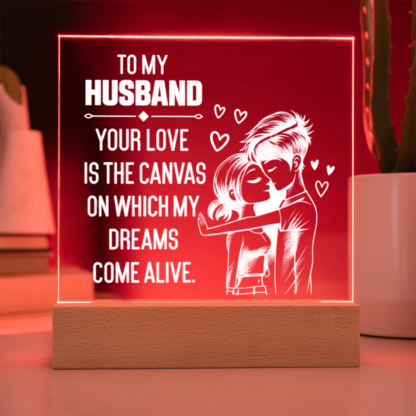 A rectangular acrylic LED light box on a wooden base, glowing red with white text that reads, 'TO MY HUSBAND YOUR LOVE IS THE CANVAS ON WHICH MY DREAMS COME ALIVE,' plus a white illustration of a couple and small hearts, shown on a tabletop in a softly blurred indoor setting.
