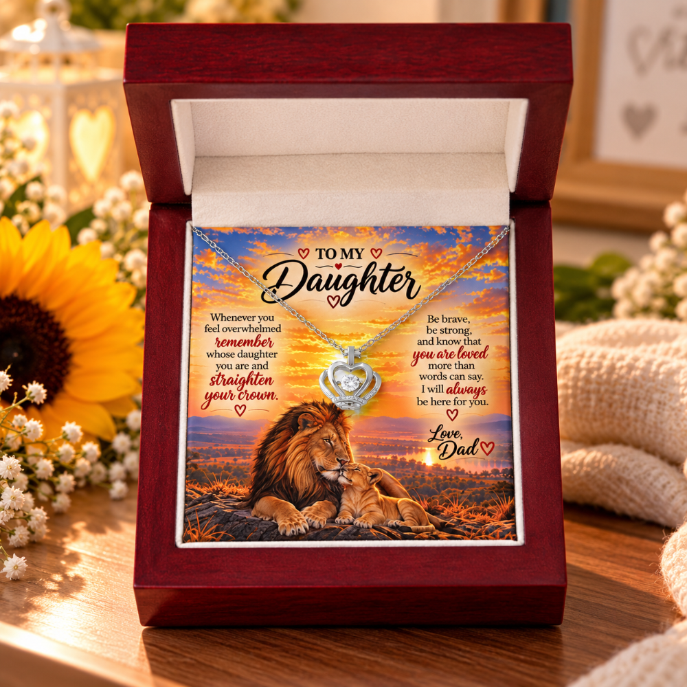 A heart-shaped pendant necklace displayed inside an open dark red wooden gift box. The pendant hangs over a printed message card addressed 'TO MY Daughter' with a lion and lioness illustration and warm sunset text.