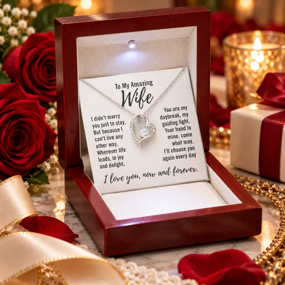 An open dark red jewelry gift box containing a heart-shaped silver pendant necklace with a clear stone, displayed on a printed message card. The box is surrounded by red roses, a lit candle, and a wrapped gift box in a romantic setting.