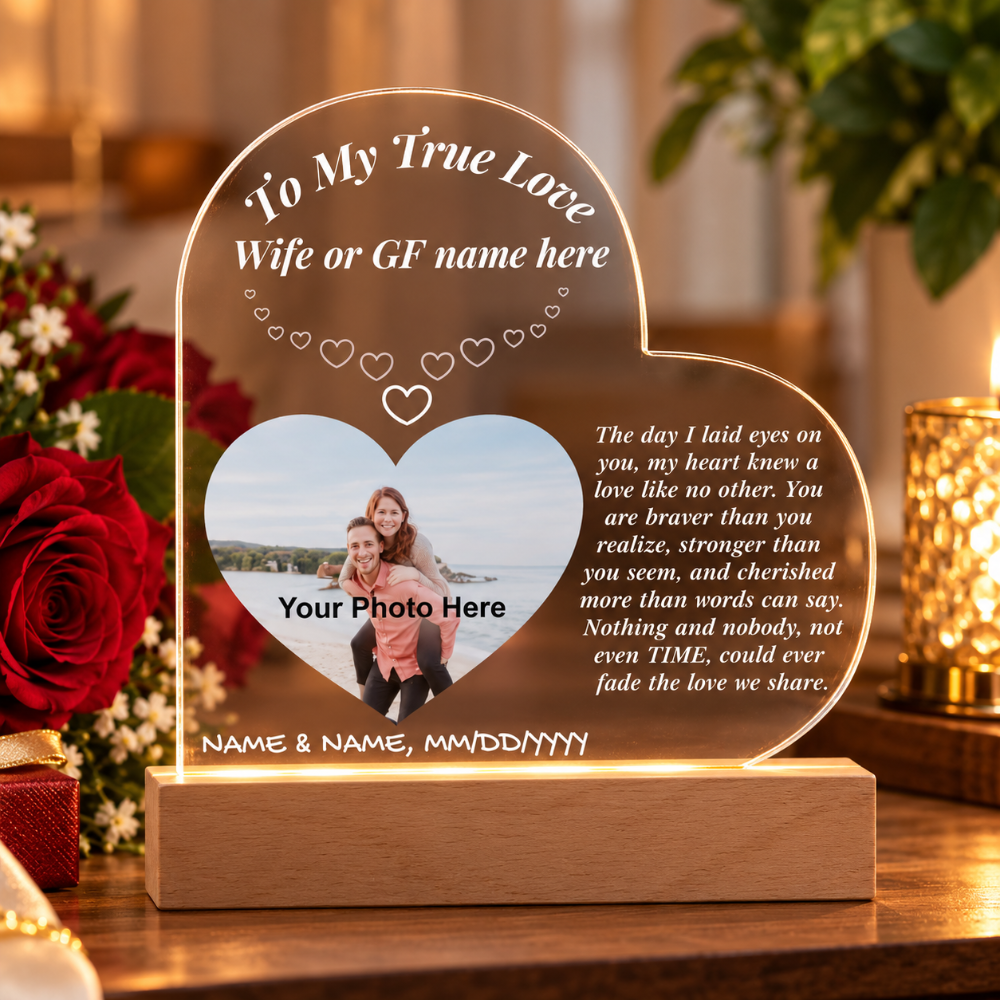 Personalized acrylic heart-shaped plaque on a wooden base, displayed on a table with blurred red roses and warm ambient lights in the background. The plaque includes customizable text and a photo slot.