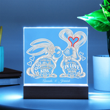 A square acrylic LED light box on a dark wooden base, glowing with a soft blue light. The front features a white engraved design of two kissing rabbits with hearts and the words 'I Still Get Butterflies Before I Kiss' and 'And I Fall In Love All Over Again,' plus the names 'Amanda & Jeremiah.