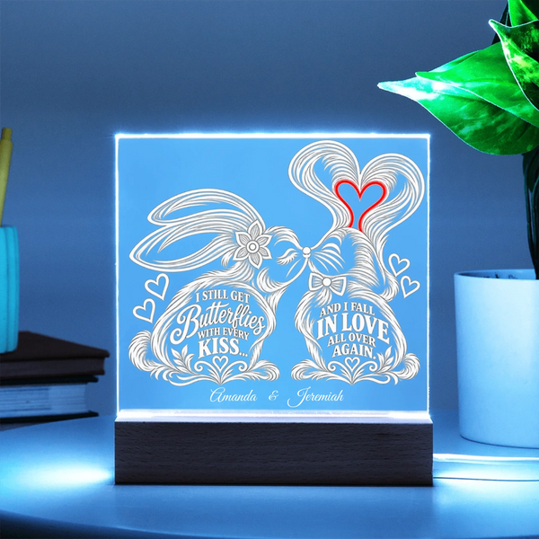 A square acrylic LED light box on a dark wooden base, glowing with a soft blue light. The front features a white engraved design of two kissing rabbits with hearts and the words 'I Still Get Butterflies Before I Kiss' and 'And I Fall In Love All Over Again,' plus the names 'Amanda & Jeremiah.