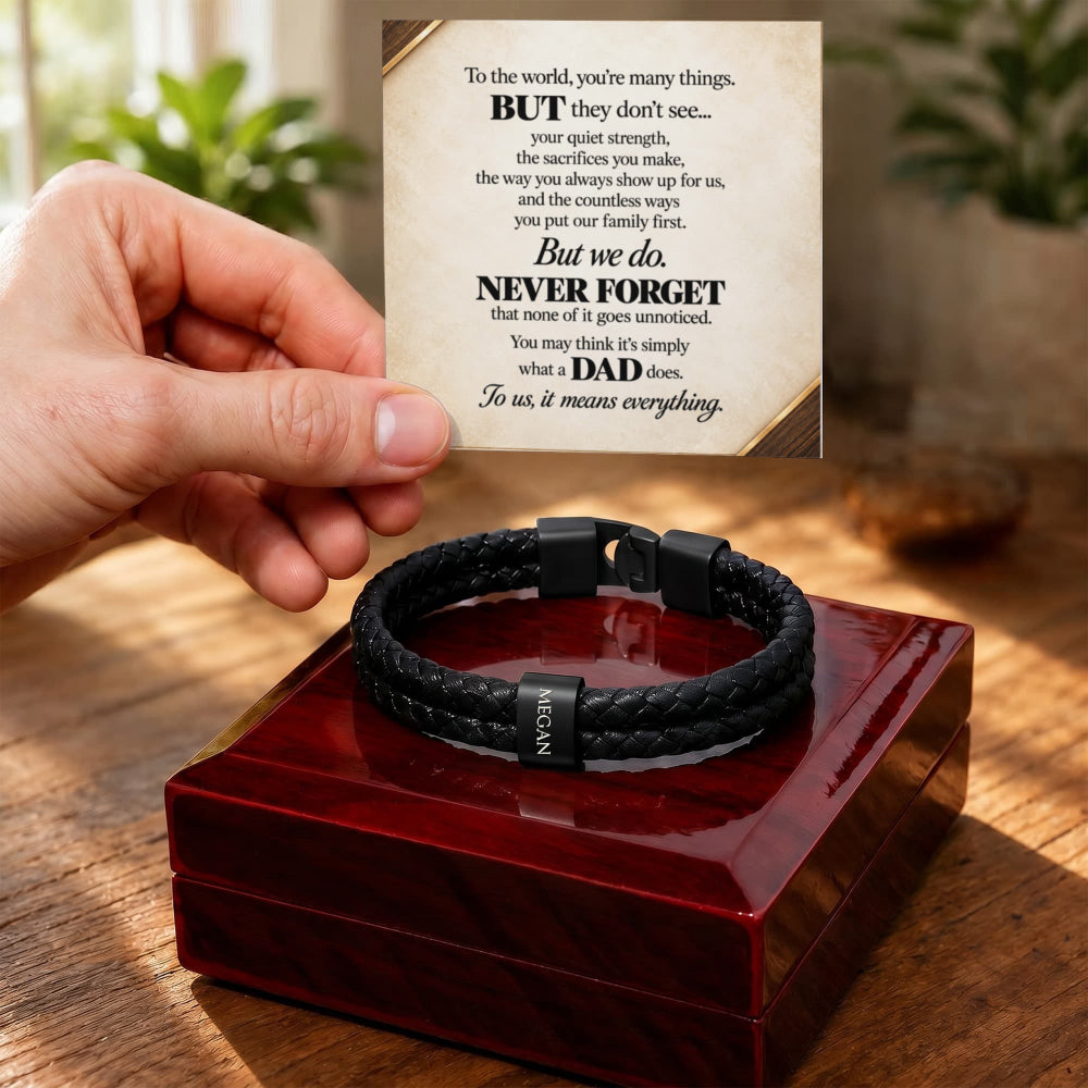 A black braided cord bracelet with a black metal clasp sits on a glossy red presentation box. A hand holds up a printed message card above the box, with a blurred indoor background behind it.