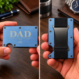 A split-screen close-up of a hand holding a slim blue metal card holder wallet. The left side shows the front with engraved gold text reading 'DAD 1st & 1982', and the right side shows the back with a black cardholder clip and black fabric loop.