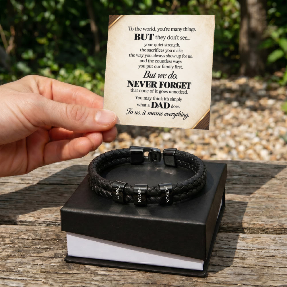 A black braided leather bracelet with silver-toned metal accents and engraved lettering, displayed on a black gift box. A hand holds a printed message card above the box, set outdoors on a wooden table with blurred greenery in the background.
