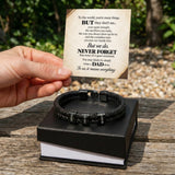 A black braided leather bracelet with silver-toned metal accents and engraved lettering, displayed on a black gift box. A hand holds a printed message card above the box, set outdoors on a wooden table with blurred greenery in the background.