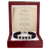 A black braided bracelet with silver letter beads displayed inside an open reddish-brown wooden gift box. The box contains a cream-colored message card with a romantic poem titled 'Two Strangers'.