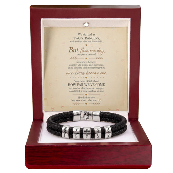 A black braided bracelet with silver letter beads displayed inside an open reddish-brown wooden gift box. The box contains a cream-colored message card with a romantic poem titled 'Two Strangers'.
