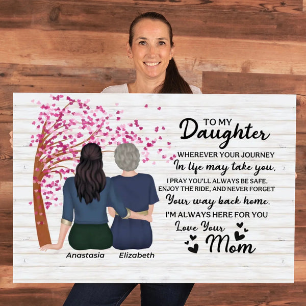 A smiling woman holds a large personalized memorial sign against a wood plank background. The sign features an illustration of two women from behind under a pink cherry blossom tree, with a heartfelt message addressed to a daughter's mom.