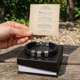 A hand holds a printed card above a black leather bracelet with silver engraved metal accents, displayed on a black gift box outdoors on a wooden table.
