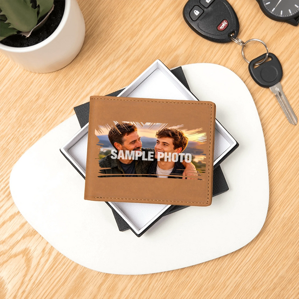A personalized brown faux-leather bifold wallet with a custom photo of two men and the text 'SAMPLE PHOTO' printed across the front, displayed on a white and black gift box on a light wood desk.