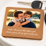 A tan leather wallet or keepsake card shown on a white textured surface with a key partly visible at the top. The wallet has a photo window displaying a man and child, overlaid with the words “SAMPLE PHOTO,” and printed text below reading, “Dad, wherever life takes you, a little piece of me goes with you.