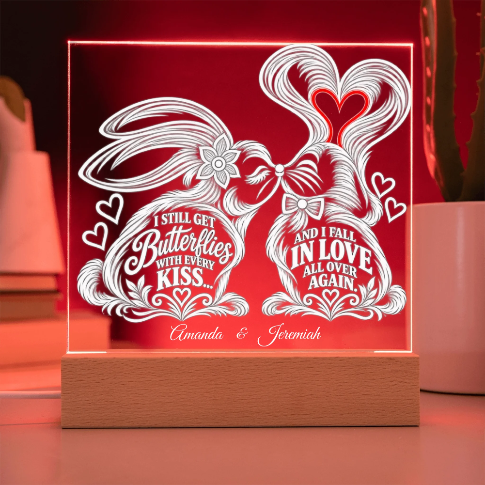 A personalized acrylic LED night light on a wooden base, glowing with a warm red light. The clear panel features white laser-cut butterfly and heart designs with the text 'I STILL GET Butterflies WITH EVERY KISS.
