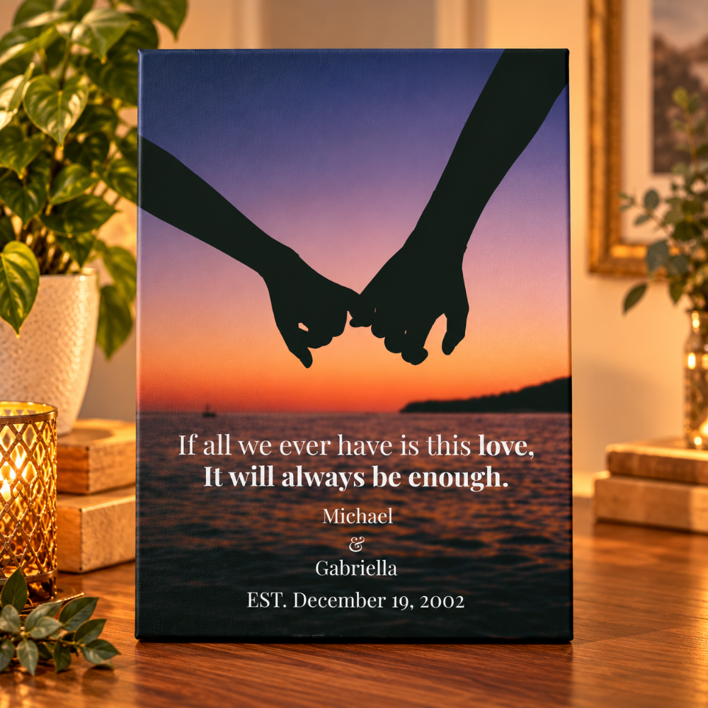 A personalized canvas print standing on a wooden table, featuring a sunset sky with two silhouetted hands holding, over water, with the text: 'If all we ever have is this love, It will always be enough. Michael & Gabriella EST.