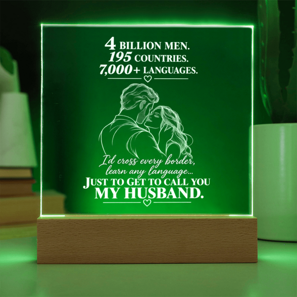 A rectangular acrylic LED light box with a wooden base, glowing green and displaying a romantic message about a husband, set against a softly blurred home background.