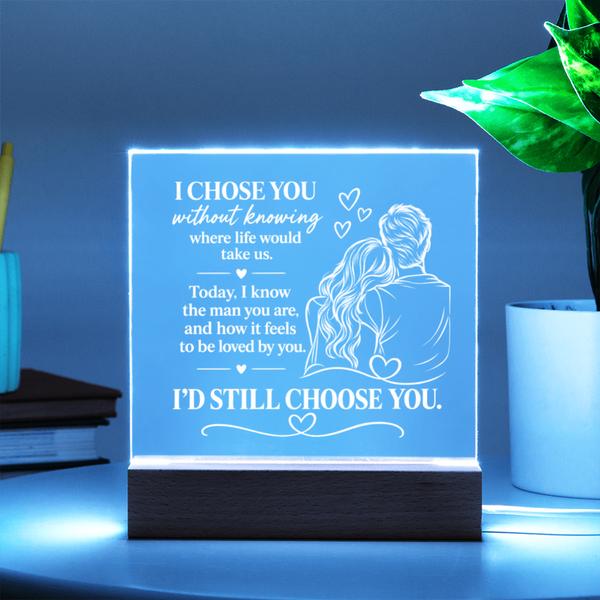 A glowing acrylic LED cube lamp on a dark wooden base, displaying a romantic quote and an illustration of a couple. The lamp is styled on a table next to a potted plant and books, with a cool blue ambient glow.