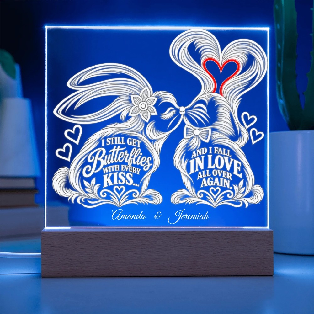 A rectangular acrylic LED light box on a wooden base, glowing with a blue border and white engraved bunny illustration and romantic quotes. The front text reads 'I STILL GET Butterflies WITH EVERY KISS.