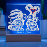 A rectangular acrylic LED light box on a wooden base, glowing with a blue border and white engraved bunny illustration and romantic quotes. The front text reads 'I STILL GET Butterflies WITH EVERY KISS.