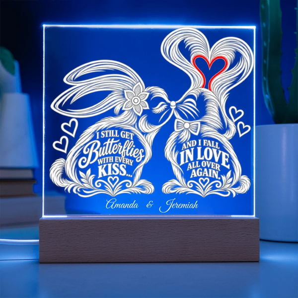 A rectangular acrylic LED light box on a wooden base, glowing with a blue border and white engraved bunny illustration and romantic quotes. The front text reads 'I STILL GET Butterflies WITH EVERY KISS.