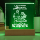 A 3D LED acrylic lamp with a wooden base, featuring a romantic quote about women and languages, with a silhouette of a couple holding hands. The lamp glows with a green light, set against a softly blurred background with a white mug.