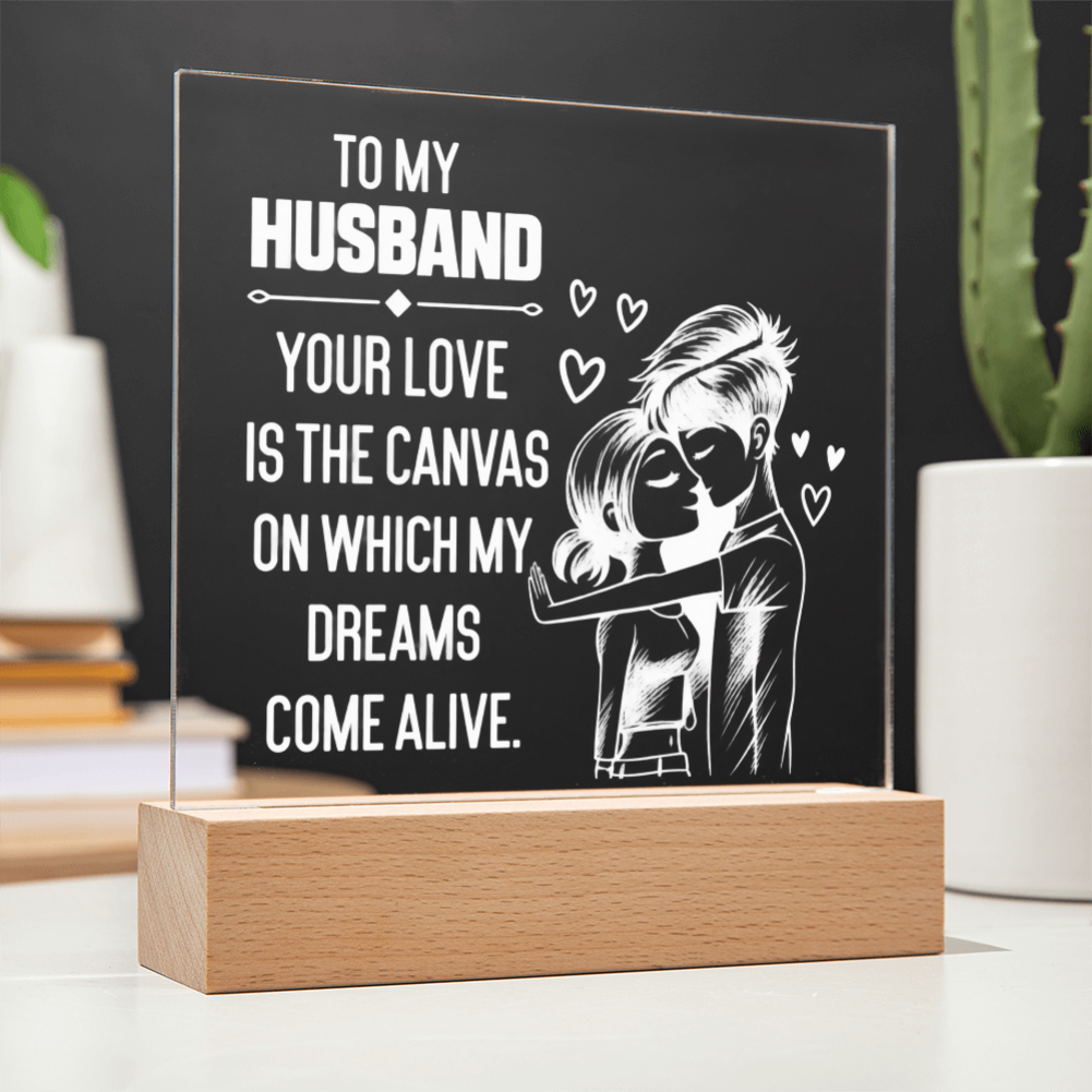 A personalized acrylic plaque on a wooden base, featuring a black background with white text that reads 'TO MY HUSBAND YOUR LOVE IS THE CANVAS ON WHICH MY DREAMS COME ALIVE.' and an illustration of a couple embracing with small hearts.