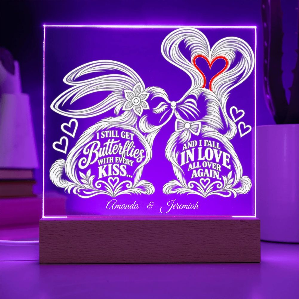 A rectangular LED acrylic light sign on a wooden base, glowing with purple light. The white design features two rabbits forming a heart shape with their tails, decorated with hearts and the words 'I STILL GET Butterflies WITH EVERY KISS.