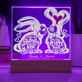 A rectangular LED acrylic light sign on a wooden base, glowing with purple light. The white design features two rabbits forming a heart shape with their tails, decorated with hearts and the words 'I STILL GET Butterflies WITH EVERY KISS.