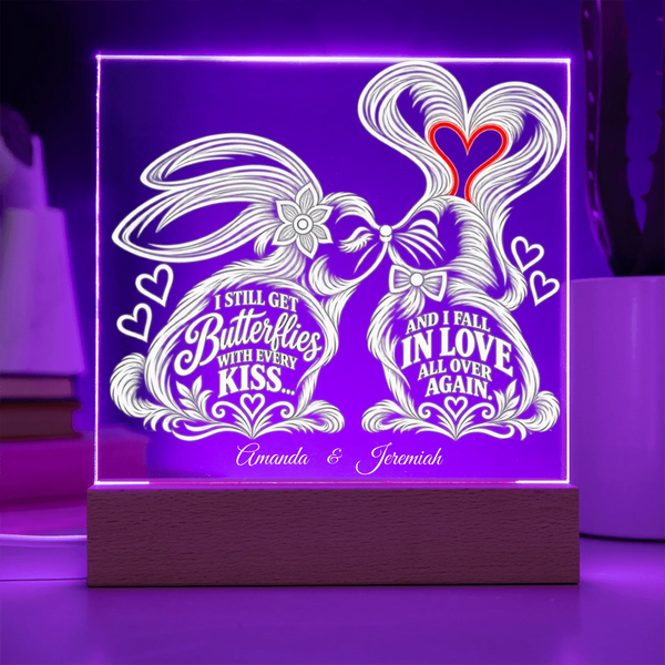 A rectangular LED acrylic light sign on a wooden base, glowing with purple light. The white design features two rabbits forming a heart shape with their tails, decorated with hearts and the words 'I STILL GET Butterflies WITH EVERY KISS.