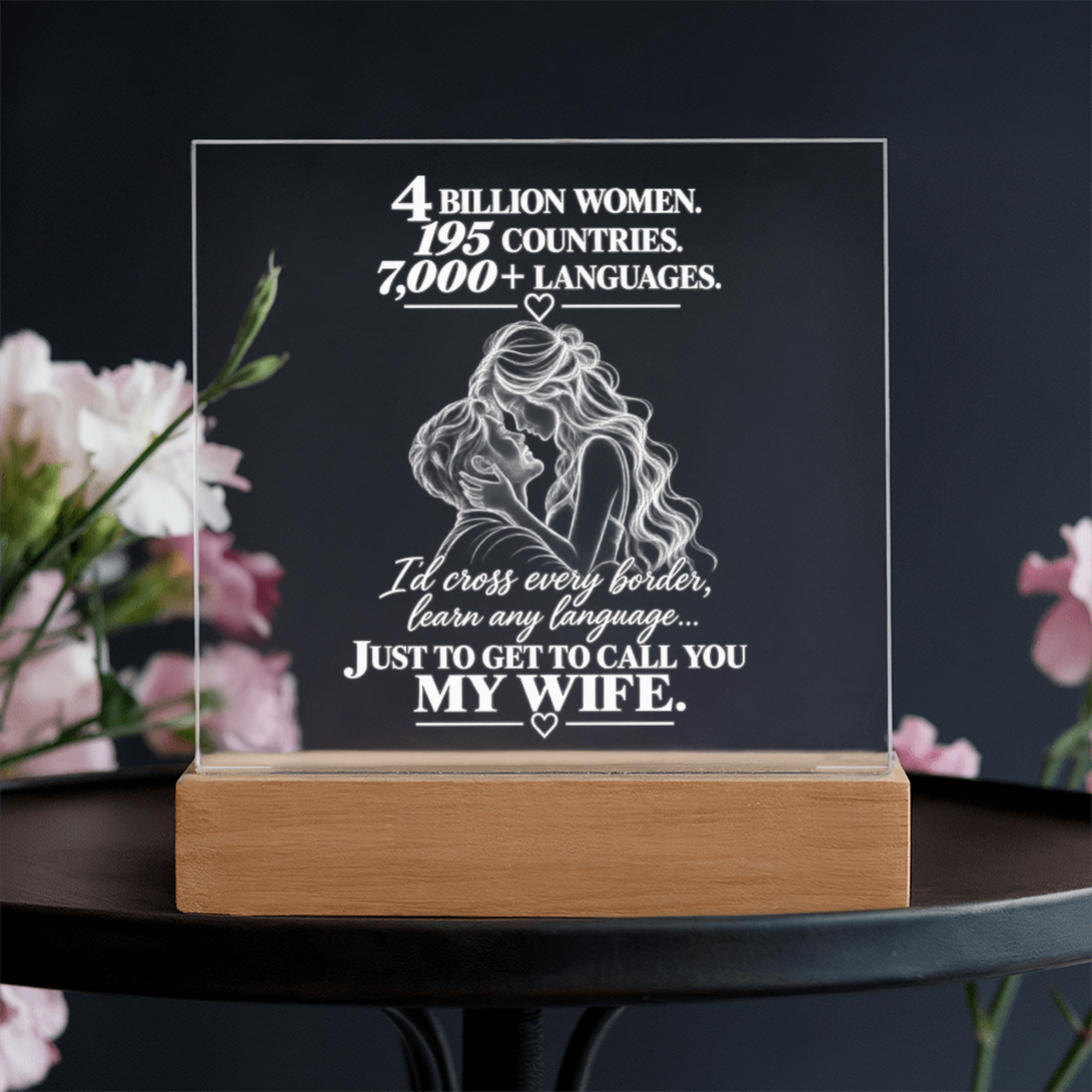 A clear acrylic plaque on a light wood base, displayed on a dark table with blurred pink flowers in the background. The plaque features white text celebrating women and a white line drawing of a couple embracing.