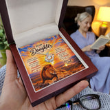 A hand holds an open dark wood keepsake box showing a custom printed card with a lion and sunset design and a silver heart-shaped pendant necklace. In the blurred background, a woman in a home setting reads a book by a lamp.