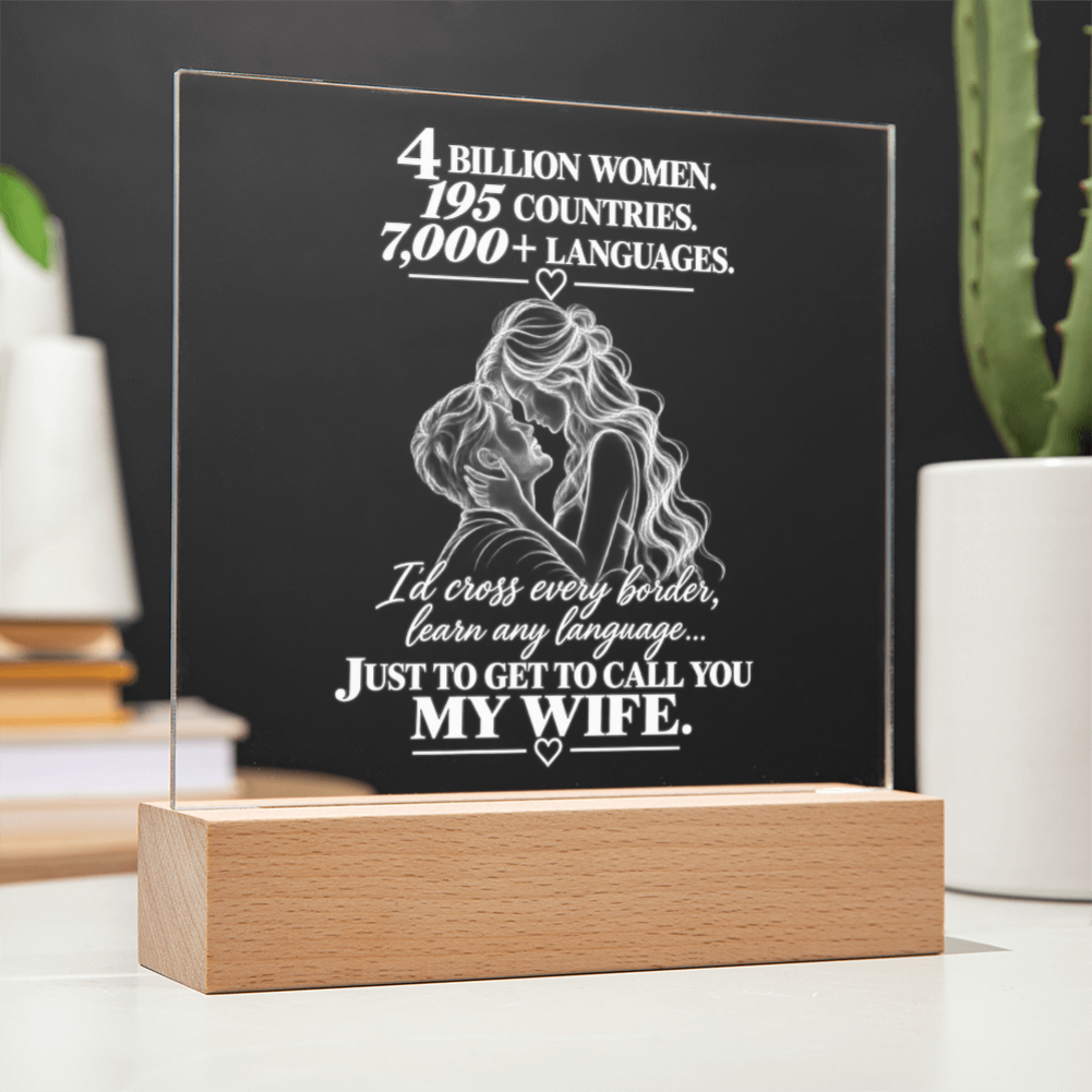 A personalized acrylic LED light plaque on a wooden block base, featuring white engraved text and an illustration of a couple, displayed on a tabletop with blurred plants and books in the background.