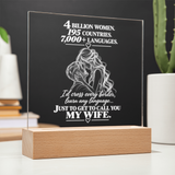 A personalized acrylic LED light plaque on a wooden block base, featuring white engraved text and an illustration of a couple, displayed on a tabletop with blurred plants and books in the background.