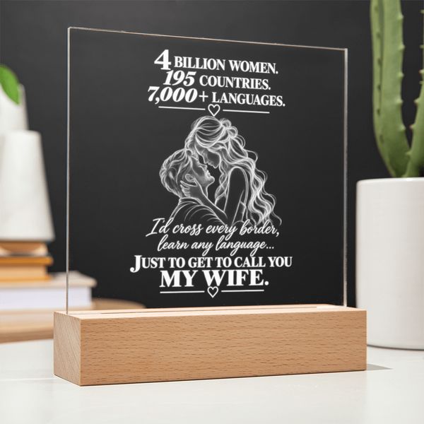 A personalized acrylic LED light plaque on a wooden block base, featuring white engraved text and an illustration of a couple, displayed on a tabletop with blurred plants and books in the background.