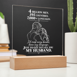 A personalized acrylic LED light plaque on a wooden base, featuring a white engraved illustration of a couple and a touching quote about love, displayed on a tabletop with blurred plants and books in the background.