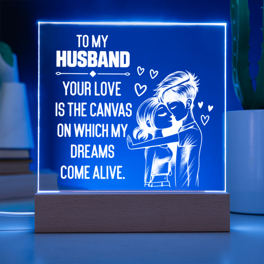 A personalized acrylic LED night light on a wooden base, glowing with a blue light. The light features white engraved text that reads 'TO MY HUSBAND YOUR LOVE IS THE CANVAS ON WHICH MY DREAMS COME ALIVE' alongside an illustration of a couple embracing with small hearts floating around.